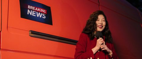 Asian female reporter smiling with microphone besides a news van at night.
