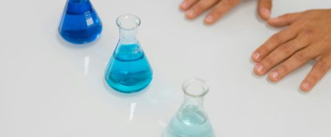 Blue liquids in Erlenmeyer flasks on a lab table with hands visible, symbolizing science research.
